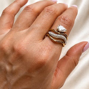 May include: A gold-toned ring featuring a multi-band design with wavy, interlocking segments. The top band is accented with a single pear-shaped clear stone, while the middle band is encrusted with a row of small, sparkling clear stones.
