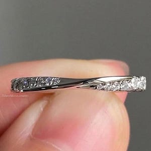 Peut inclure: Une bague argentée présentant un design de bande torsadée avec une série de petites pierres claires et scintillantes incrustées le long de la surface. Le métal a une finition polie et réfléchissante, et la bande se courbe légèrement au centre.