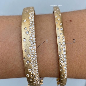 May include: Two gold-toned bangles with a brushed matte finish, each featuring a cluster of embedded clear stones that transition from dense to sparse. The wider bangle is labeled 1 and the narrower bangle is labeled 2, with SilverMuseJewels text.