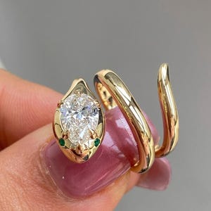 Può includere: Un anello in oro a forma di serpente con una pietra preziosa trasparente taglio a pera come testa, due piccole pietre verdi come occhi e una fascia liscia e lucida che si avvolge attorno al dito. Il testo SilverMuseJewels è visibile sull'anello.