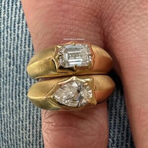 Peut inclure: Deux bagues en or superposées avec une finition mate brossée. La bague supérieure présente un diamant taille émeraude rectangulaire et la bague inférieure un diamant en forme de poire. Les pierres sont serties. Le texte SilverMuseJewels est visible.