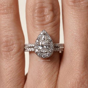 Peut inclure: Une bague argentée avec une pierre centrale en forme de poire entourée d'un halo de petites pierres rondes. L'anneau est incrusté d'une rangée de petites pierres rondes. Le texte SilverMuseJewels est visible à la base du sertissage.