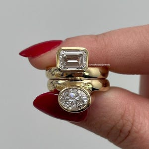 May include: Two stacked gold rings featuring bezel-set stones. The top ring has a rectangular emerald-cut clear stone, and the bottom ring has an oval-cut clear stone. The text SilverMuseJewels is visible on the side of the top ring band.