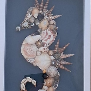 May include: A framed wall art piece featuring a seahorse mosaic crafted from various natural seashells in shades of white, tan, and brown. The seahorse is set against a dark gray background within a white frame, with the initials HLO in the corner.