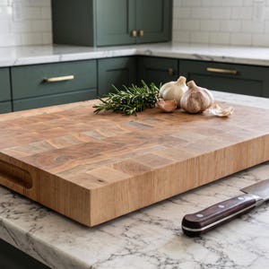 May include: A thick rectangular wooden end-grain chopping board with integrated side handles and a circular brand logo on the right edge. The board features a natural light brown wood grain pattern and sits on a white marble countertop.