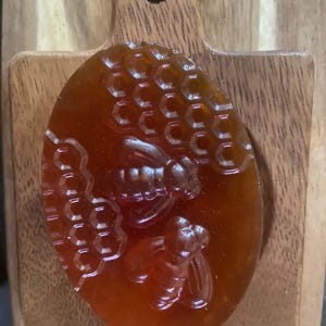 Puede incluir: Una pastilla de jabón ovalada y translúcida de color ámbar con un diseño en relieve de dos abejas y un patrón de panal en su superficie. El jabón tiene una textura suave y brillante con un tono profundo similar a la miel.