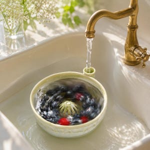 May include: A light green ceramic berry colander with a central drainage cone and a side spout, positioned under a brass faucet. The bowl contains a swirling mixture of fresh blueberries and red raspberries being rinsed with running water.