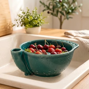 May include: A deep teal ceramic berry colander with a rounded bowl shape, small drainage holes, and a side handle with integrated holes. The colander is filled with fresh red strawberries and dark cherries, with water droplets visible on the surface.