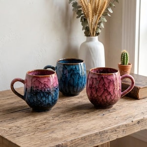 Puede incluir: Tres tazas de cerámica con cuerpos redondeados y asas. Una taza presenta un degradado de rosa y azul, otra es de color azul profundo con acabado brillante, y la tercera es de color rosa y marrón oscuro con un patrón texturizado de escamas.