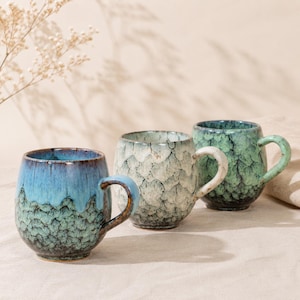 May include: Three ceramic mugs with rounded bodies and handles. The first mug features a blue and green drip glaze. The second mug has a cream and grey crackle pattern. The third mug displays a deep green and black textured glaze finish.