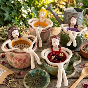 Op de afbeelding: Vier keramische thee-infusers in de vorm van figuren die in kommen rusten. Elk figuur heeft geschilderd haar en kleding in oranje, paars of rood. De kommen hebben bloemmotieven en groene of roze glazuren. Eén kom bevat rode vruchten en witte bloemen.