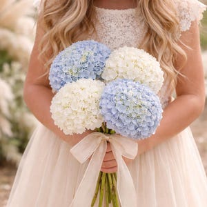 May include: A bouquet of white and pale blue hydrangeas tied with a cream ribbon. The flowers are held in front of a cream-coloured dress.