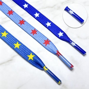 May include: Three flat shoelaces with distinct patterns. One is dark blue with yellow stars, one is light blue with red stars, and one is dark blue with white stars. Each lace features a matching plastic aglet at the end for easy threading.