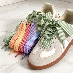 May include: A pair of off-white sneakers with gum soles, featuring wide green gingham ribbon laces tied in a bow. Beside the sneakers are five additional pairs of flat ribbon shoelaces in green, orange, yellow, red, purple, and blue gingham patterns.