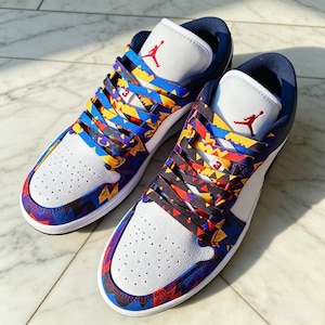 May include: A pair of low-top sneakers featuring white perforated leather toe boxes, dark blue collars, and white tongues with red Jumpman logos. The overlays and laces display a vibrant geometric pattern in shades of blue, yellow, and red.