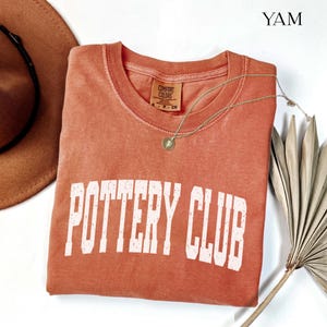 May include: A terracotta-colored short-sleeve t-shirt with the text POTTERY CLUB printed in white block letters across the chest. The shirt features a Comfort Colors brand label at the inner neckline and is labeled with the color name YAM.