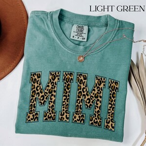 Könnte beinhalten: Ein hellgrünes Kurzarm-T-Shirt mit Rundhalsausschnitt und einem Comfort Colors Markenetikett. Auf der Vorderseite steht das Wort MIMI in großen Blockbuchstaben mit einem braun-schwarzen Leopardenmuster. Eine dünne Goldkette liegt auf dem T-Shirt.