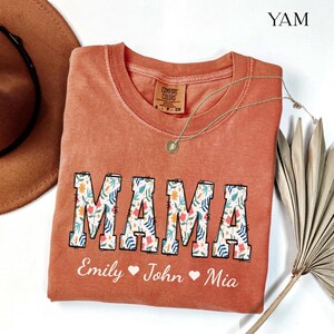 以下が含まれることがあります： テラコッタカラーの半袖Tシャツ。花と葉の模様で埋め尽くされた大きなブロック体の「MAMA」という文字がプリントされています。その下には、白い筆記体でEmily、John、Miaという名前とハートのアイコンが描かれています。
