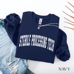 Könnte beinhalten: Ein marineblaues Sweatshirt mit Rundhalsausschnitt und dem weißen Schriftzug STERILE PROCESSING TECH in einer abgenutzten, gebogenen Schriftart auf der Brust. Das Kleidungsstück hat eine silberfarbene Kette am Kragen und ein Gildan-Etikett.