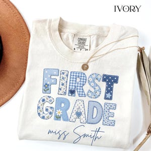 May include: An ivory t-shirt with the text FIRST GRADE in blue patterned block letters featuring floral, gingham, and polka dot designs. Below the text, the name miss Smith is printed in blue script. A gold necklace rests on the neckline.