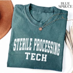 Könnte beinhalten: Ein blau-fichtenfarbenes T-Shirt mit dem weißen Schriftzug STERILE PROCESSING TECH in einer abgenutzten Schriftart. Das Shirt hat einen Rundhalsausschnitt und wird mit einer goldfarbenen Halskette auf dem Stoff präsentiert.