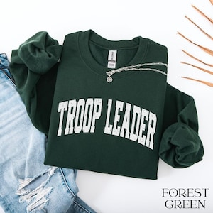 May include: A forest green crewneck sweatshirt featuring the text TROOP LEADER in a distressed white collegiate font across the chest. The garment includes a silver chain necklace resting on the collar and is paired with light blue distressed denim.