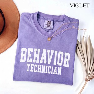 May include: A violet short-sleeved t-shirt with a crew neckline featuring the white distressed text "BEHAVIOR TECHNICIAN" printed on the front. The shirt has a Comfort Colors brand label inside the collar and a thin gold-toned necklace.