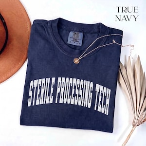 May include: A navy blue short-sleeve t-shirt with a crew neckline and the text STERILE PROCESSING TECH printed in white across the chest. The shirt features a Comfort Colors brand label at the inner neck and is labeled as size S.