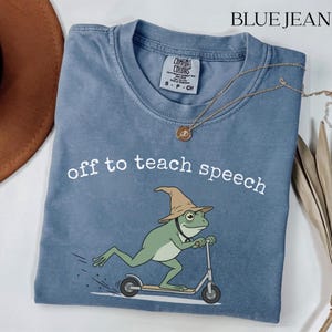 Op de afbeelding: Een blauw spijkerkleurig t-shirt met een afbeelding van een groene kikker met een beige tovenaarshoed op een zilveren step. De tekst "off to teach speech" staat in het wit boven de kikker gedrukt. Het shirt heeft een Comfort Colors label.