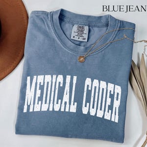 Może przedstawiać: Krótkorękawowa koszulka w kolorze dżinsowego błękitu z okrągłym dekoltem. Na piersi znajduje się biały napis MEDICAL CODER wykonany przecieraną, łukowatą czcionką. Wewnątrz przy kołnierzu widoczna jest metka marki Comfort Colors.