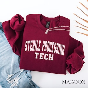 May include: A maroon crewneck sweatshirt featuring the white distressed text STERILE PROCESSING TECH printed across the chest. The garment is displayed alongside blue denim jeans and a delicate silver chain necklace.