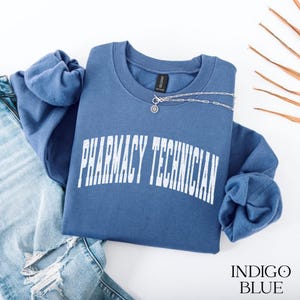 May include: An indigo blue crewneck sweatshirt featuring the white text PHARMACY TECHNICIAN in a distressed, arched font across the chest. The garment is paired with distressed blue denim jeans and a silver chain necklace.