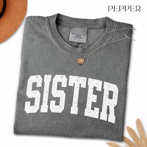 May include: A charcoal grey short-sleeve t-shirt with a distressed white graphic print that reads SISTER in a bold, collegiate-style font. The shirt features a crew neckline and a garment-dyed finish, with the word PEPPER printed in the top right corner.