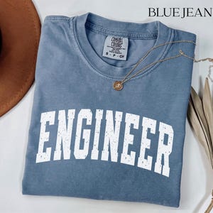 May include: A blue jean colored short-sleeve t-shirt with a crew neckline. The shirt features the word ENGINEER printed in large, distressed white block letters across the chest. A gold-toned necklace rests on the fabric near the collar.