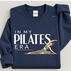 May include: A navy blue crewneck sweatshirt featuring the white text "IN MY PILATES ERA" and a graphic of a person in olive green activewear performing a side-stretching yoga pose on a mat. The inner neck label reads "softubee".
