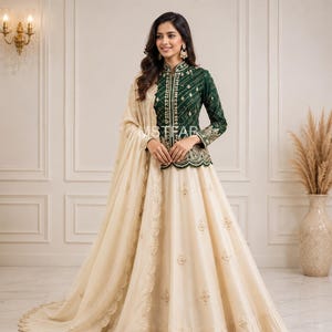 Könnte beinhalten: Eine dunkelgrüne langärmelige Jacke mit Goldstickerei und Stehkragen, kombiniert mit einem bodenlangen cremefarbenen Rock mit verstreuten goldenen Blumenmotiven und einem passenden cremefarbenen Dupatta mit gebogtem Stickrand.