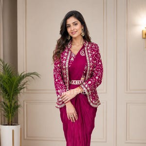 Puede incluir: Un conjunto de falda drapeada y blusa en color magenta, combinado con una chaqueta de manga larga del mismo tono con bordados florales blancos en solapas y puños, junto a un cinturón fino con hebilla metálica en la cintura.