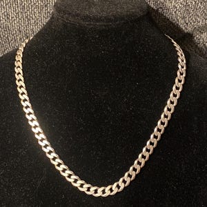 May include: A silver-toned curb chain necklace featuring interlocking, flattened oval links. The metal chain has a polished, reflective finish and is displayed in a circular shape, showcasing the uniform pattern of the connected metal links.