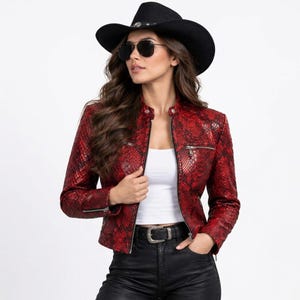 Op de afbeelding: Een jas met een rood en zwart slangenleermotief, een opstaande kraag, lange mouwen en zilverkleurige zakken met rits. De jas wordt gedragen over een effen wit shirt en gecombineerd met een zwarte broek met een zilverkleurige gesp.