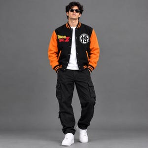 Op de afbeelding: Een zwart met oranje varsity-jack met het Dragon Ball Z-logo op de rechterborst en een wit cirkelvormig kanji-symbool op de linkerborst, gedragen over een wit t-shirt met een zwarte cargobroek en witte sneakers, gecombineerd met een zonnebril.