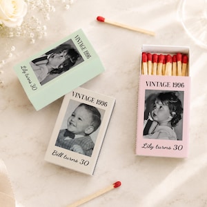May include: Three rectangular matchboxes in light green, white, and light pink, each featuring a black and white childhood photograph, the text VINTAGE 1996, and a name followed by turns 30. One pink box is open, revealing wooden matches with red tips.