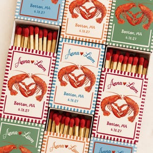 May include: Several rectangular matchboxes featuring a lobster illustration forming a heart shape, with the text Anna and Liam, Boston, MA, and 6.18.27. Each box contains wooden matches with red tips, arranged in a grid pattern with various colored borders.