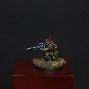 May include: A miniature soldier figure in a kneeling position, wearing green camouflage clothing, a red headband, and carrying a large rifle with a scope. The figure is mounted on a circular base covered in textured brown grass-like material.
