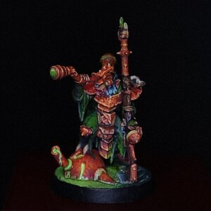 Puede incluir: Una figura en miniatura pintada de una criatura parecida a un roedor con armadura naranja y verde. El personaje sostiene un bastón largo en una mano y un telescopio en la otra, sobre una base naranja redondeada con detalles en verde brillante.