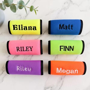 May include: Six rectangular neoprene handle covers with black trim, each featuring a personalised name embroidered in black or white thread. The colours include neon yellow, blue, pink, lime green, purple, and orange, displaying the names Eliana, Matt, RILEY, FINN, Riley, and Megan.