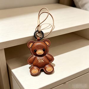 以下が含まれることがあります： 茶色の型押しレザーで作られたクマの形をしたキーホルダー。ベージュの鼻と足の裏、黒い目があしらわれています。クマの胸の中央には「RK」の文字が刻印されています。黒い丸いリングと細いベージュのコードが付いています。
