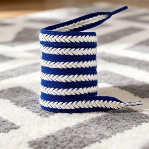 Puede incluir: Un cordón plano con una base de color azul real vibrante y un patrón trenzado o de espiga blanco prominente que recorre el centro. El cordón está enrollado en forma cilíndrica e incluye herretes de plástico azul sólido en ambos extremos.