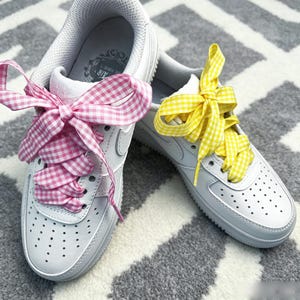 May include: A pair of white sneakers featuring perforated toe boxes and side panels. One sneaker is laced with wide pink and white gingham ribbon laces tied in a large bow, while the other features yellow and white gingham ribbon laces.