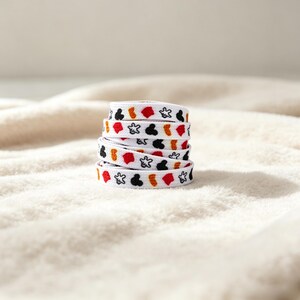 May include: A white flat shoelace coiled into a stack, featuring a repeating pattern of black mouse head silhouettes, white glove outlines, red shapes, and orange textured spots printed along the entire length of the fabric material.