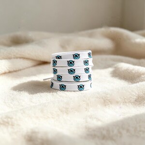 May include: A white shoelace coiled into a stack, featuring a repeating pattern of small, light blue circular faces wearing black sunglasses. The shoelace is flat and woven, with the graphic print evenly spaced along the entire length of the fabric.